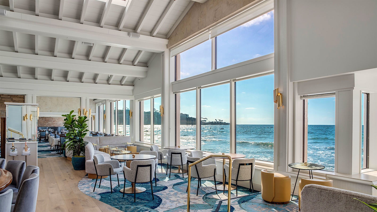 The dining room at the Marine Room, with a tall white ceiling, windows looking out on to the ocean, white cloth chairs and a blue, modern design carpet